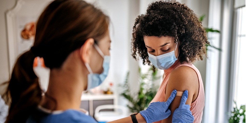 A person in scrubs and a facemask applies a bandage to a person's arm. 